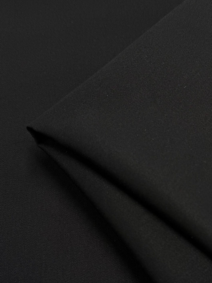 The Gabardine - Black - 150cm by Super Cheap Fabrics showcases a smooth texture with fine diagonal twill lines. Draped into a neat, gentle pointed fold, the durable and slightly reflective fabric gives off an elegant appearance.