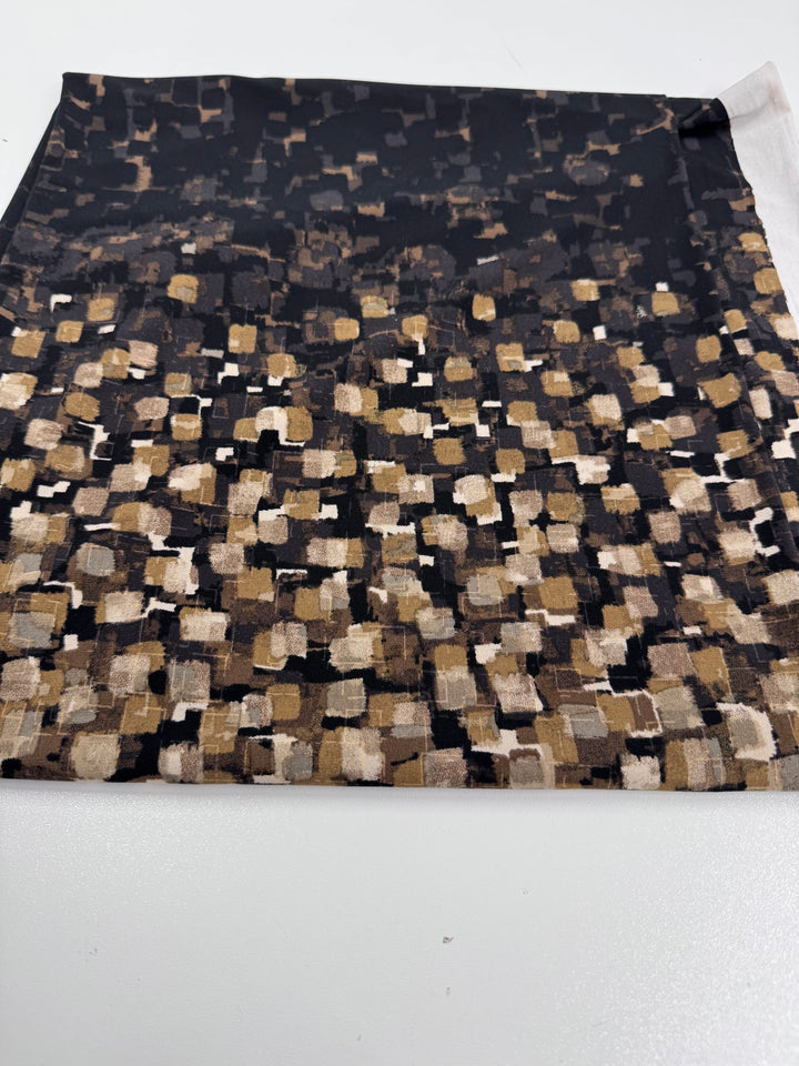The Designer Lycra Panel - 90cm Panel from Super Cheap Fabrics features small, irregular gold, brown, cream, and black squares in a gradient on a dark base. It’s neatly folded on a white surface to show its full width and panel length.