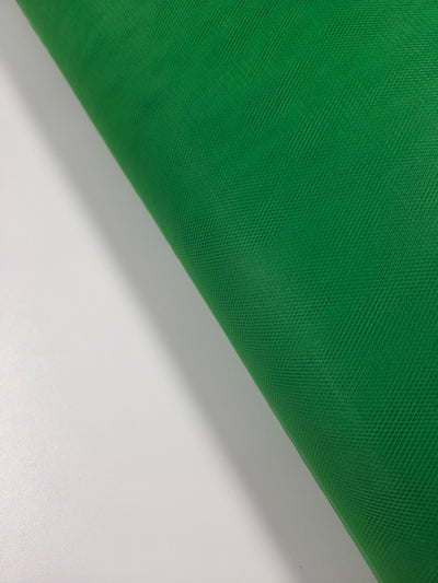 A close-up of Super Cheap Fabrics' Soft Tulle - Emerald - 150cm shows a diagonally placed roll of green, lightweight, textured fabric on a white background.