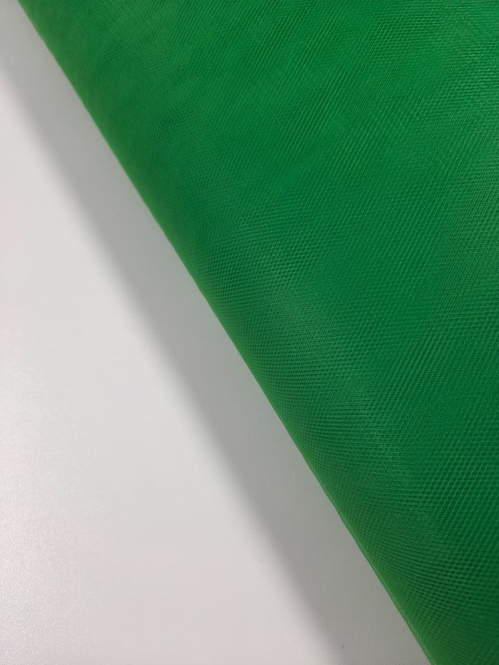 A close-up of Super Cheap Fabrics' Soft Tulle - Emerald - 150cm shows a diagonally placed roll of green, lightweight, textured fabric on a white background.