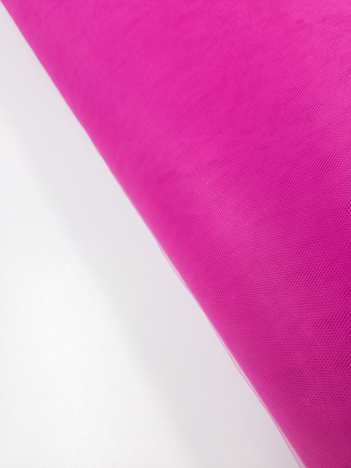 A close-up of Soft Tulle - Very Berry (150cm) by Super Cheap Fabrics, partially unrolled on a white surface. This sheer, lightweight, and slightly textured bright pink fabric is ideal for bridal veils or costumes.