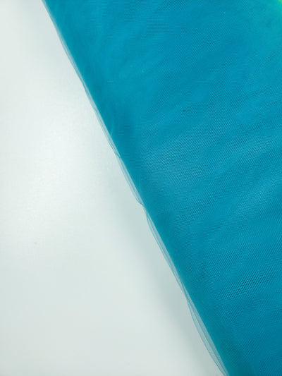 A roll of Soft Tulle - Lake Blue - 150cm from Super Cheap Fabrics lies diagonally on a white surface—ideal for bridal veils or craft projects.