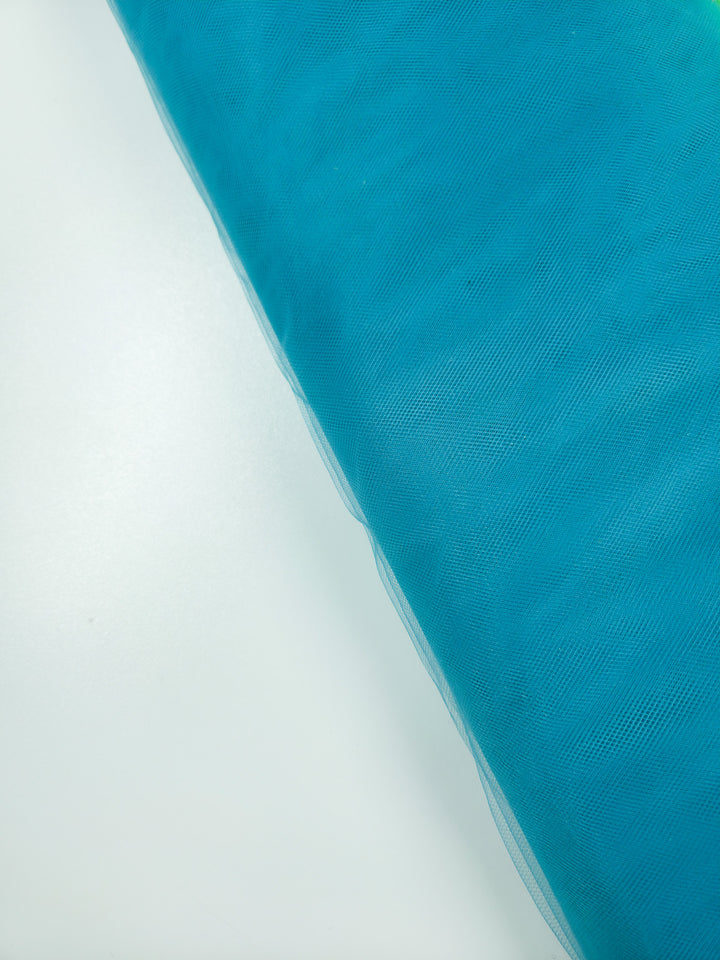 A roll of Soft Tulle - Lake Blue - 150cm from Super Cheap Fabrics lies diagonally on a white surface—ideal for bridal veils or craft projects.