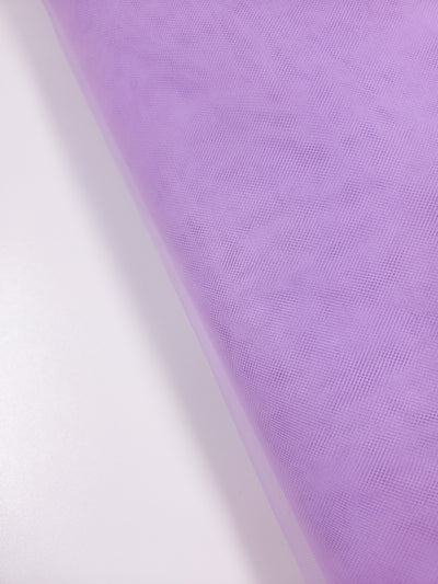 A close-up of Super Cheap Fabrics’ Soft Tulle – Lavender – 150cm shows its sheer, delicate texture and fine mesh, ideal for bridal veils or crafts. The roll is diagonally placed on a smooth white surface.