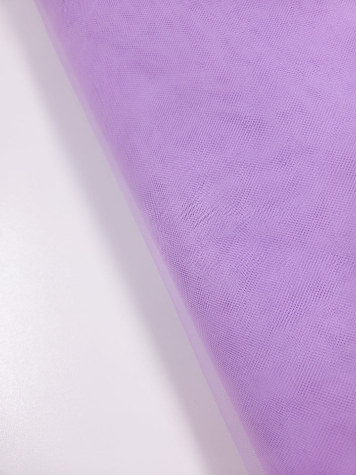 A close-up of Super Cheap Fabrics’ Soft Tulle – Lavender – 150cm shows its sheer, delicate texture and fine mesh, ideal for bridal veils or crafts. The roll is diagonally placed on a smooth white surface.