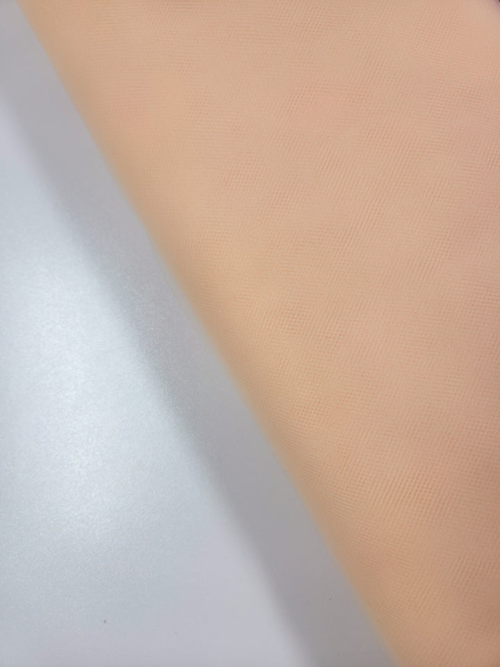A close-up of Super Cheap Fabrics’ Soft Tulle - Peach - 150cm on a white surface highlights its soft, slightly textured look—ideal for sheer, lightweight projects like bridal veils.