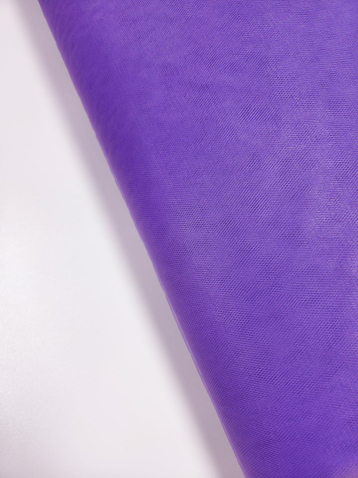 A close-up of Super Cheap Fabrics’ Soft Tulle - Purple - 150cm, partially unrolled on a white surface—ideal for creating lightweight projects like bridal veils.