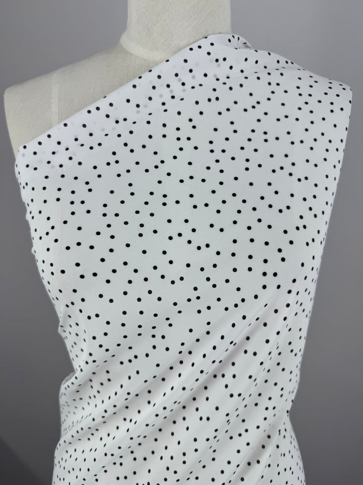 The Super Cheap Fabrics Printed Chiffon - Polka Dots - 145cm, a delicate white fabric with black dots, elegantly drapes over a mannequin's shoulder. Set against a plain gray backdrop, the lightweight material stands out beautifully, making it ideal for sewing projects.