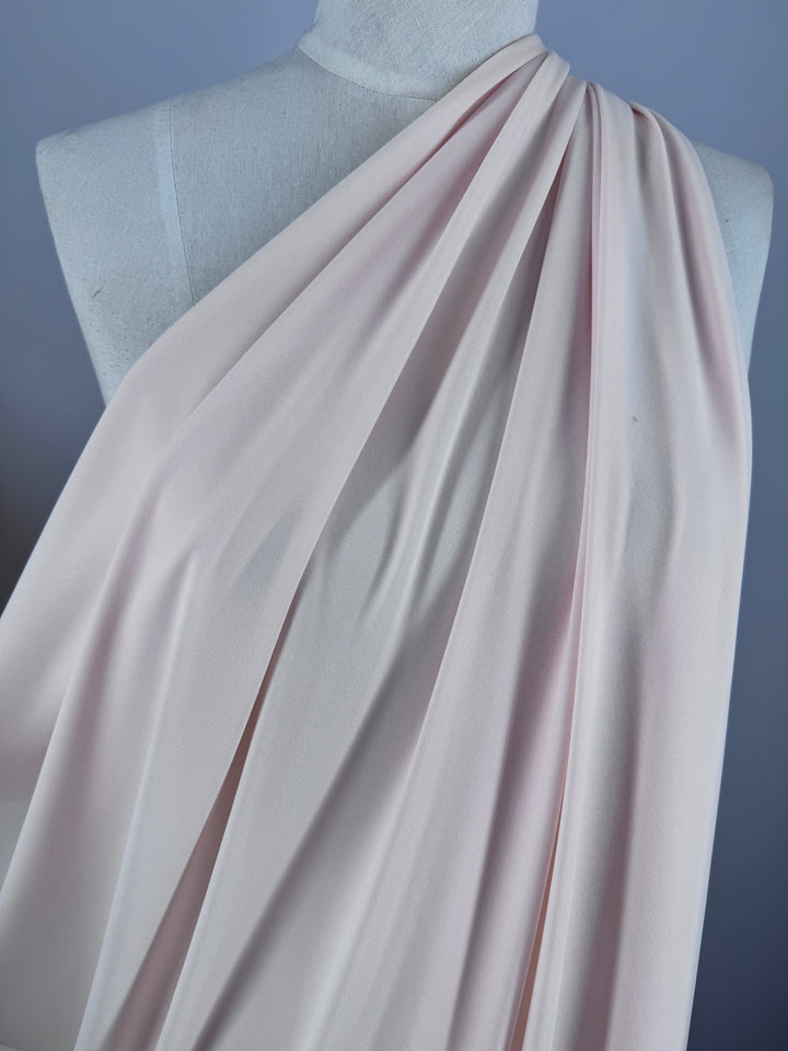 A white mannequin displays Super Cheap Fabrics’ ITY Knit in Baby Pink (150cm), elegantly pleated over one shoulder, set against a light purple background.