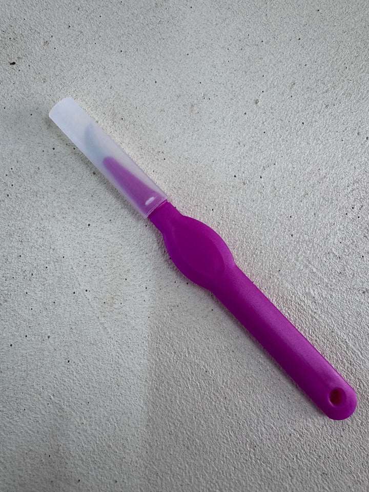 A pink seam ripper from Super Cheap Fabrics, resembling a precision thread cutter, rests on a light gray surface. The handle is pink, complementing the white blade cover.