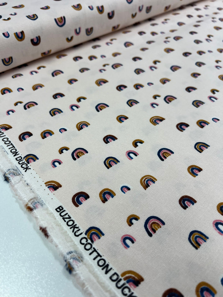 The "Printed Canvas - Wanderlust - 112cm" from Super Cheap Fabrics features small, colorful rainbows on light pink. Ideal for tote bags or home décor accents, it ensures quality and style with its Buzoku Cotton Duck label along the edge.