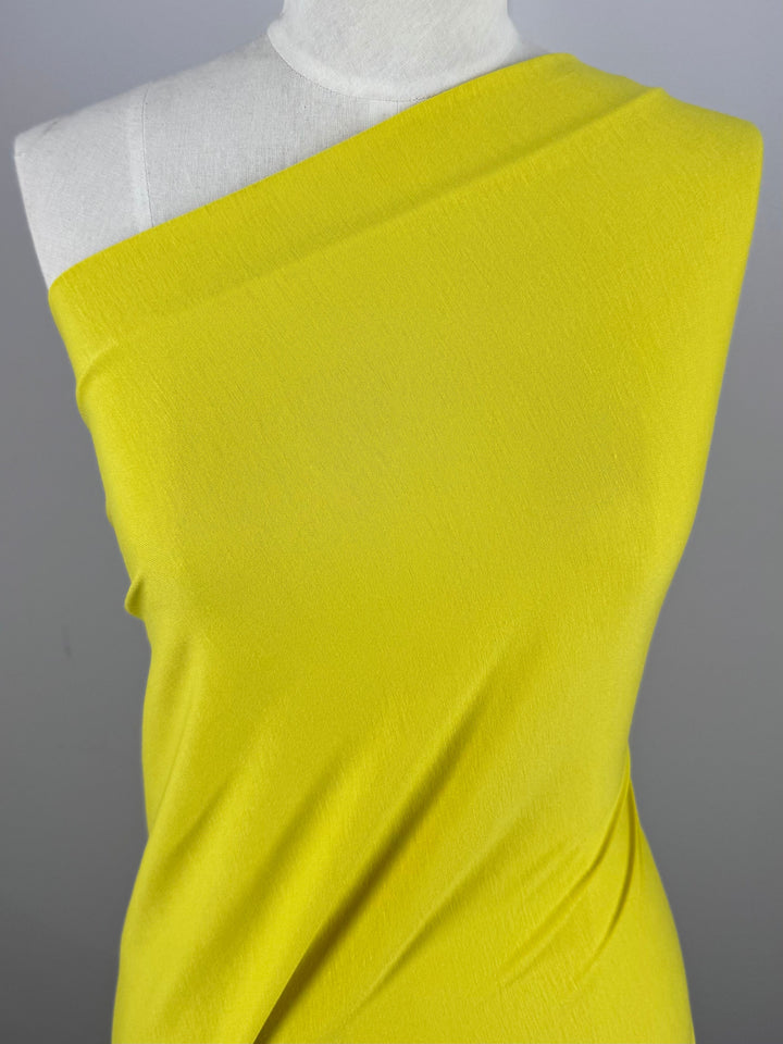 The Cotton Lycra - Green Sheen - 170cm by Super Cheap Fabrics is displayed on a mannequin with a one-shoulder design against a gray background, highlighting its medium weight and smooth, slightly stretchy texture.