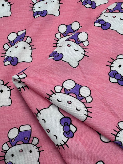 The Super Cheap Fabrics Printed Jersey - Kitty (162cm) is a lightweight poly/cotton fabric adorned with charming cartoon characters, including a white cat with a purple bow and hat, closed eyes, whiskers, and arrives neatly folded for your next creative project.