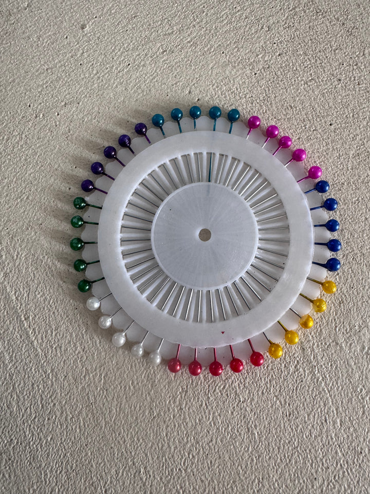 The Super Cheap Fabrics Coloured Pinwheel boasts a round design with multicolored pins in vivid shades of green, red, white, yellow, blue, and pink on a textured beige surface.