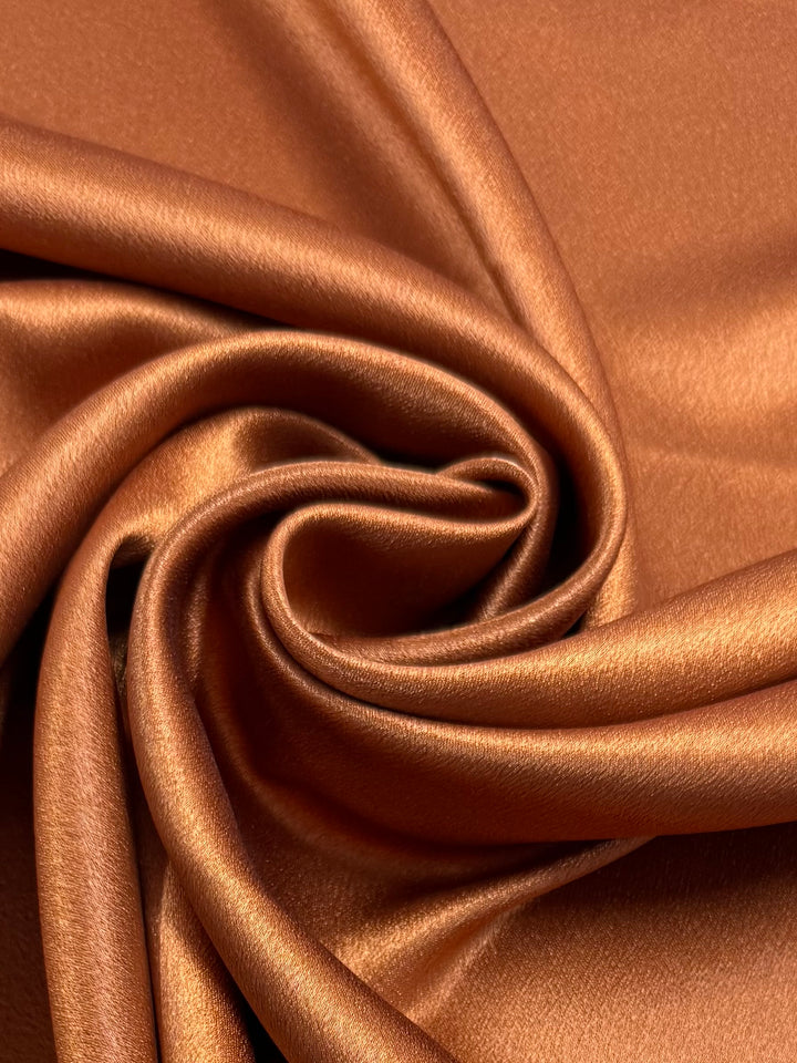 Satin Back Crepe - Desert Sun by Super Cheap Fabrics features glossy, coppery folds that form a spiral pattern, making this 150cm wide fabric ideal for elegant tops or luxurious eveningwear.