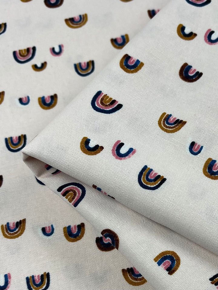 Close-up of Super Cheap Fabrics' "Printed Canvas - Wanderlust - 112cm" displaying small multicolored rainbow patterns in brown, pink, blue, and black. Ideal for home décor or crafting tote bags, the folded fabric emphasizes its vibrant print.