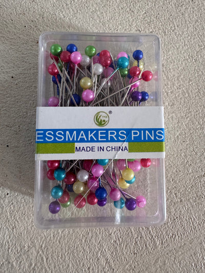 A clear plastic box of Super Cheap Fabrics Dressmakers Pins - Multicolour, Pack of 100, with round heads and a "DRESSMAKERS PINS MADE IN CHINA" label, shown on a textured light surface.