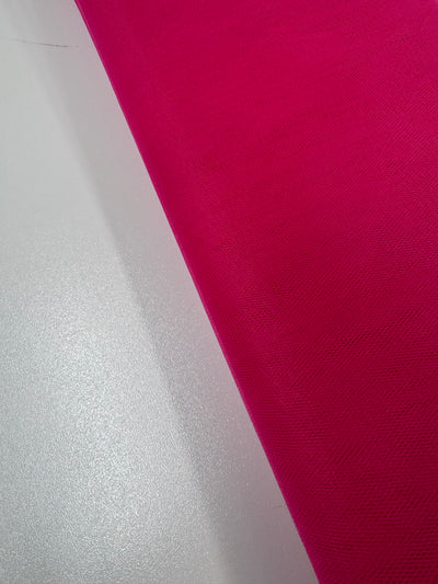A close-up shows a smooth white surface beside Super Cheap Fabrics’ Soft Tulle - Teaberry - 150cm, a bright pink, textured fabric often used in crafts and veils, with a diagonal line dividing the contrasting colors and textures.