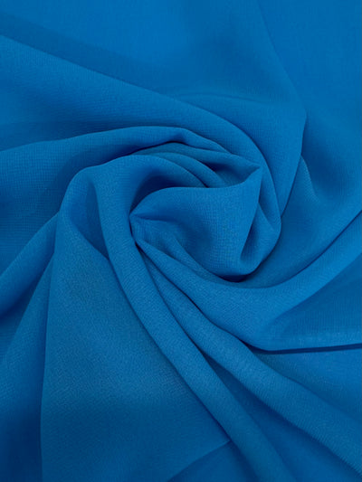A close-up of Super Cheap Fabrics' Hi-Multi Chiffon - Vivid Blue (148cm) shows vibrant blue polyester arranged in soft, swirling folds with a textured, flowing pattern.