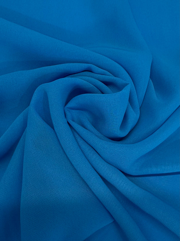 A close-up of Super Cheap Fabrics' Hi-Multi Chiffon - Vivid Blue (148cm) shows vibrant blue polyester arranged in soft, swirling folds with a textured, flowing pattern.