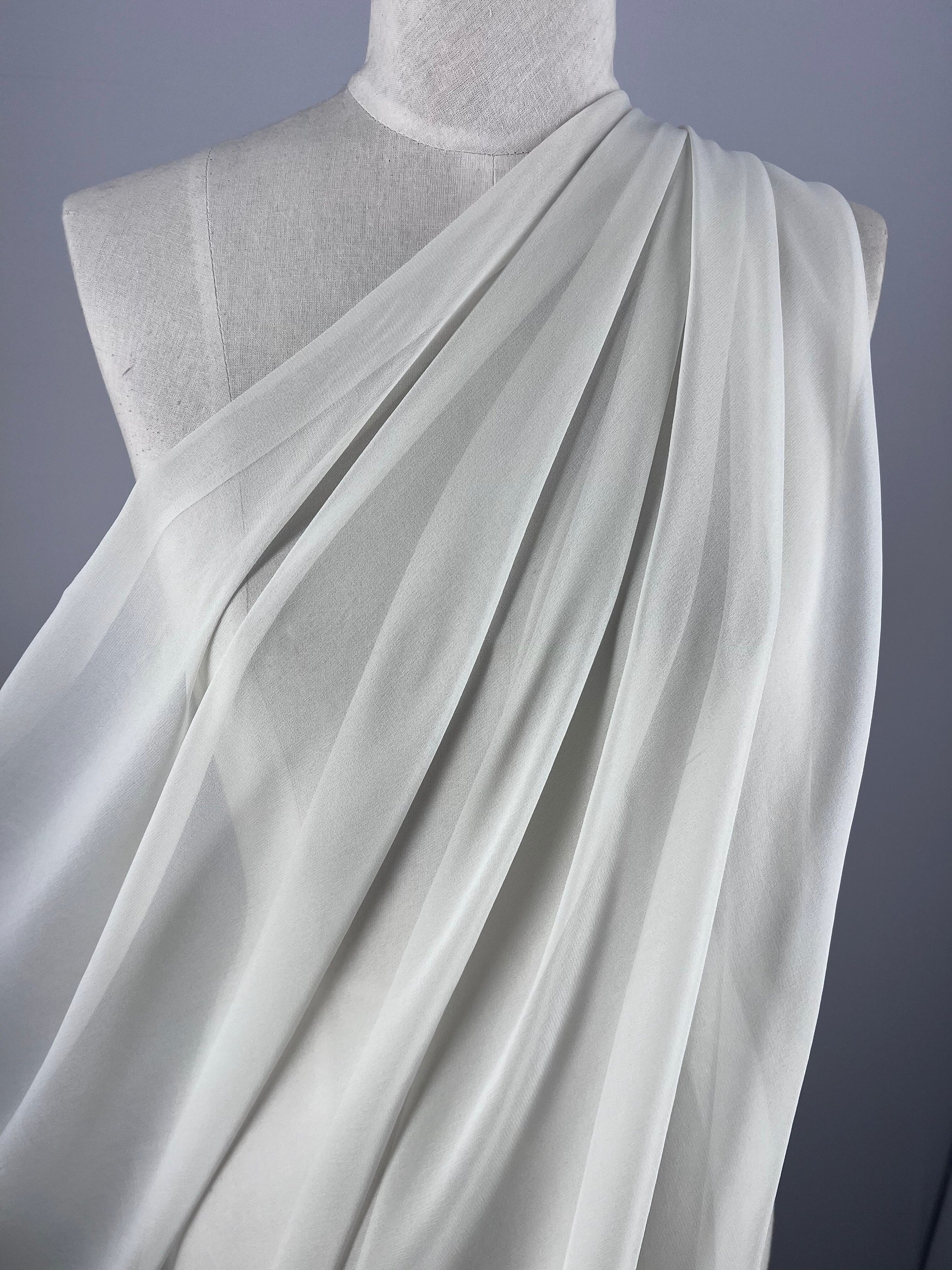 The mannequin displays Super Cheap Fabrics' Pure Silk Georgette in Silver Blue, perfect for spring and summer outfits. This lightweight, sheer fabric features an elegant asymmetrical drape with gentle folds and cascading lines on a simple gray backdrop. Width: 135cm.