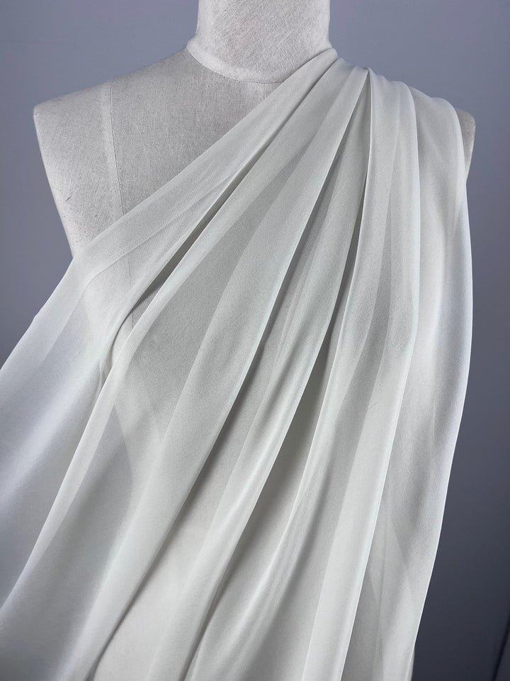 The mannequin displays Super Cheap Fabrics' Pure Silk Georgette in Silver Blue, perfect for spring and summer outfits. This lightweight, sheer fabric features an elegant asymmetrical drape with gentle folds and cascading lines on a simple gray backdrop. Width: 135cm.