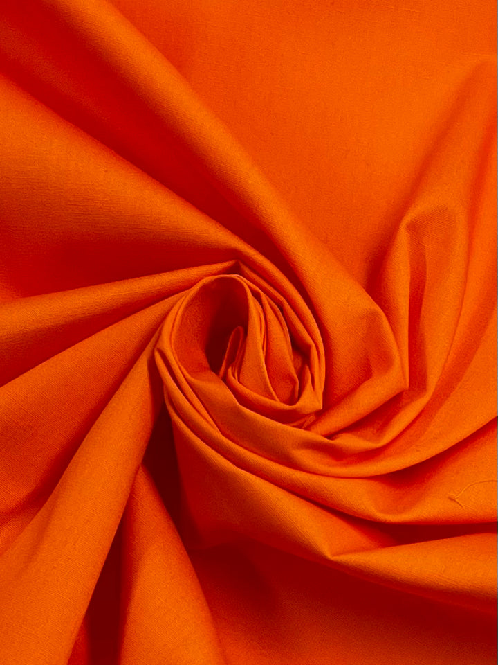 A close-up of Super Cheap Fabrics’ Plain Cotton - Orange - 150cm shows the vibrant fabric gathered in a spiral, forming soft folds and dynamic shadows.