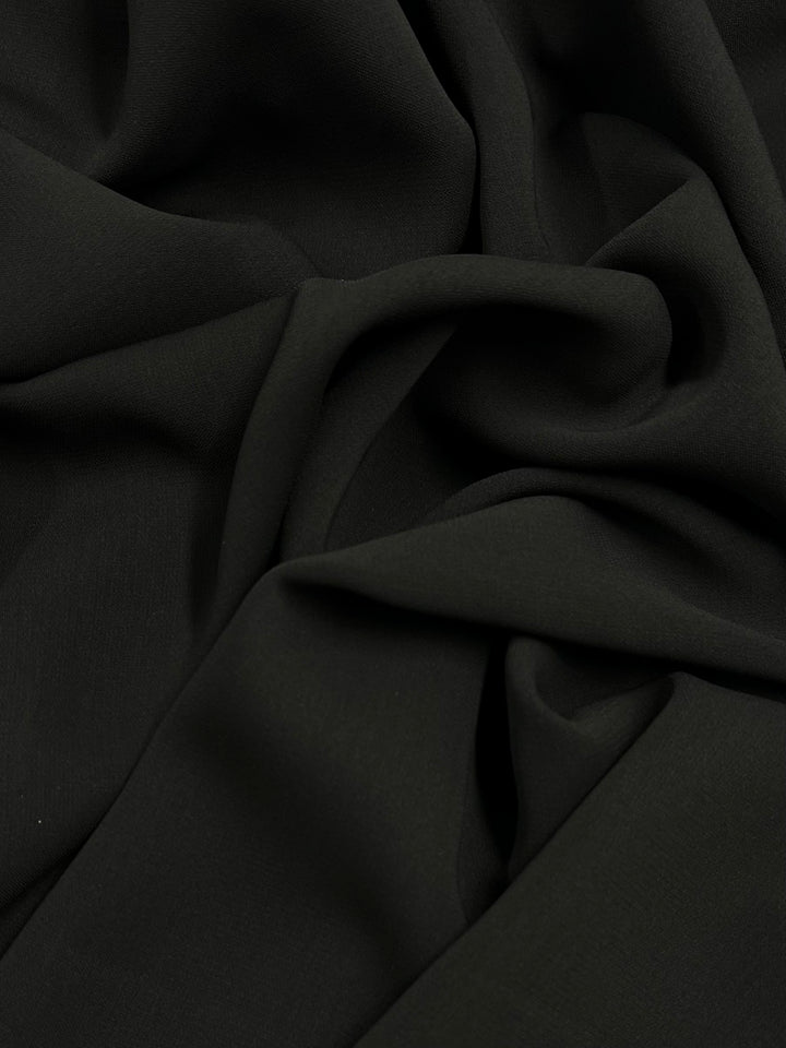 A close-up of medium weight black polyester fabric, displaying smooth folds and creases. The material appears soft and slightly shiny, with the light subtly highlighting the textures and depth of the fabric. This is Super Cheap Fabrics' Microfibre Suiting - Black - 150cm.
