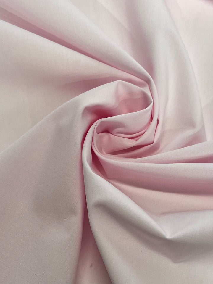 A close-up of Super Cheap Fabrics' Plain Cotton - Pink A Boo (158cm) shows soft, light pink cotton fabric draped in swirling folds and gentle shadows.
