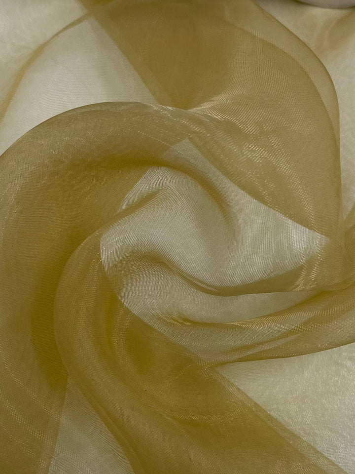 Here's a rephrased version using the given product data:

A close-up of the "Organza - Sand - 150cm" from Super Cheap Fabrics reveals its sheer, golden texture with a smooth, flowing form. This polyester material gently swirls into soft and elegant patterns while light and shadows dance across its delicate surface.
