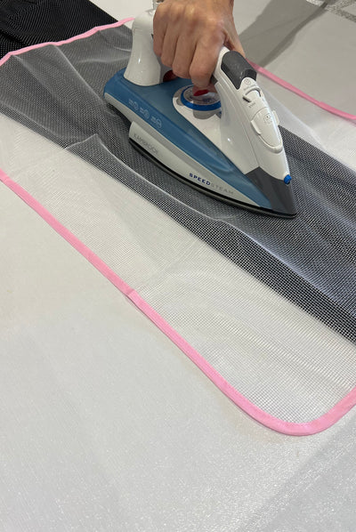 Using the Ironing Protective Cloth - Mesh by Super Cheap Fabrics, a person carefully irons mesh fabric with a pink border using a blue and white hot iron on an ironing board, focusing on its intricate texture while indoors, demonstrating how to shield clothing from direct heat.
