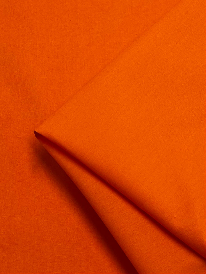 The Plain Cotton - Orange - 150cm from Super Cheap Fabrics is a lightweight, vibrant orange fabric with a smooth texture, ideal for cheerful and breezy projects. One corner is folded diagonally to showcase its quality and color.