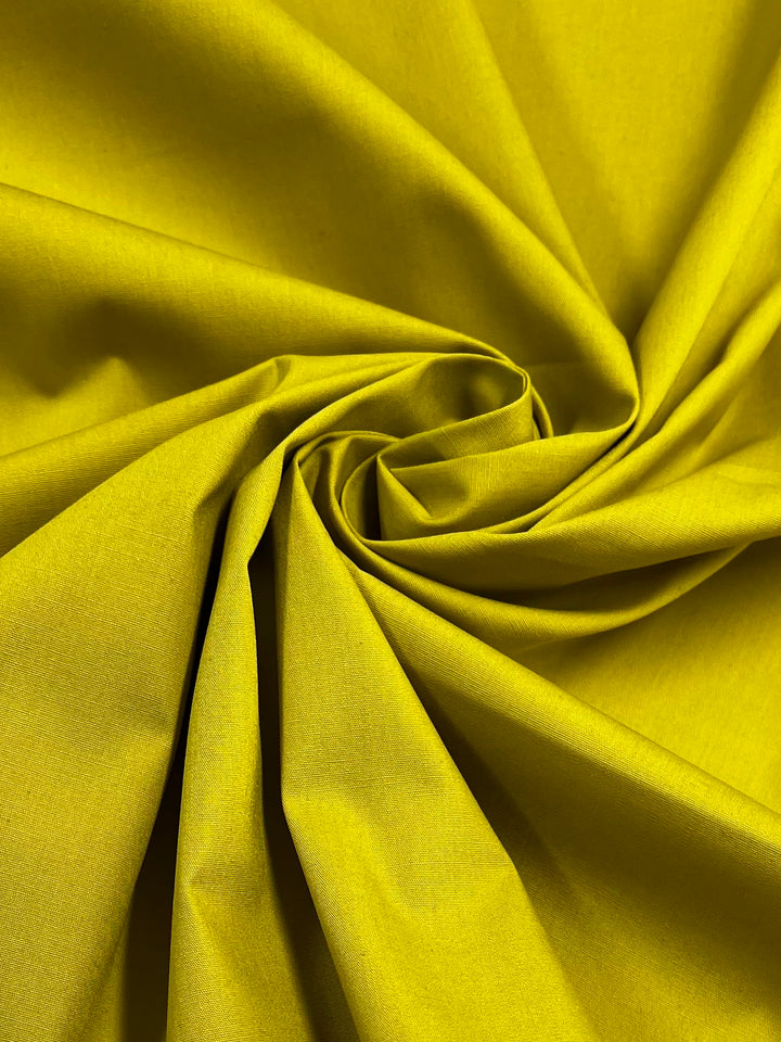 Close-up of Super Cheap Fabrics' Plain Cotton - Antique Moss (150cm), lightweight 100% cotton, gathered in a spiral to showcase its smooth texture, bright yellow hue, and elegant folds.