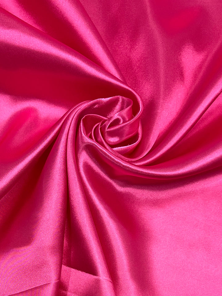 A close-up image of the smooth, shiny "Satin - Hot Pink - 150cm" fabric from Super Cheap Fabrics is arranged in a spiral fold to showcase its lustrous texture and vibrant color. Light reflections on the surface accentuate the softness and richness of this glamorous satin, making it perfect for elegant dresses or sophisticated accessories.