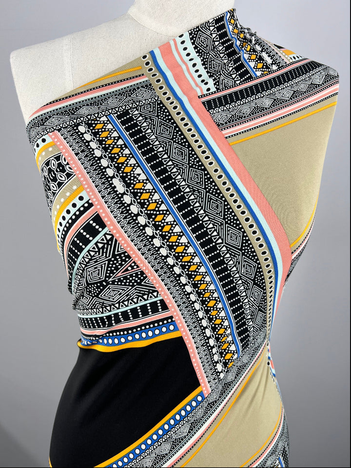 A mannequin showcases fabric from Super Cheap Fabrics, featuring their REMNANT - Lycra - Aztec Wrap in a colorful display. The piece spans 160cm and boasts an intricate geometric pattern with stripes, dots, and abstract shapes in shades of black, white, blue, pink, yellow, and beige on an 80cm wide panel.