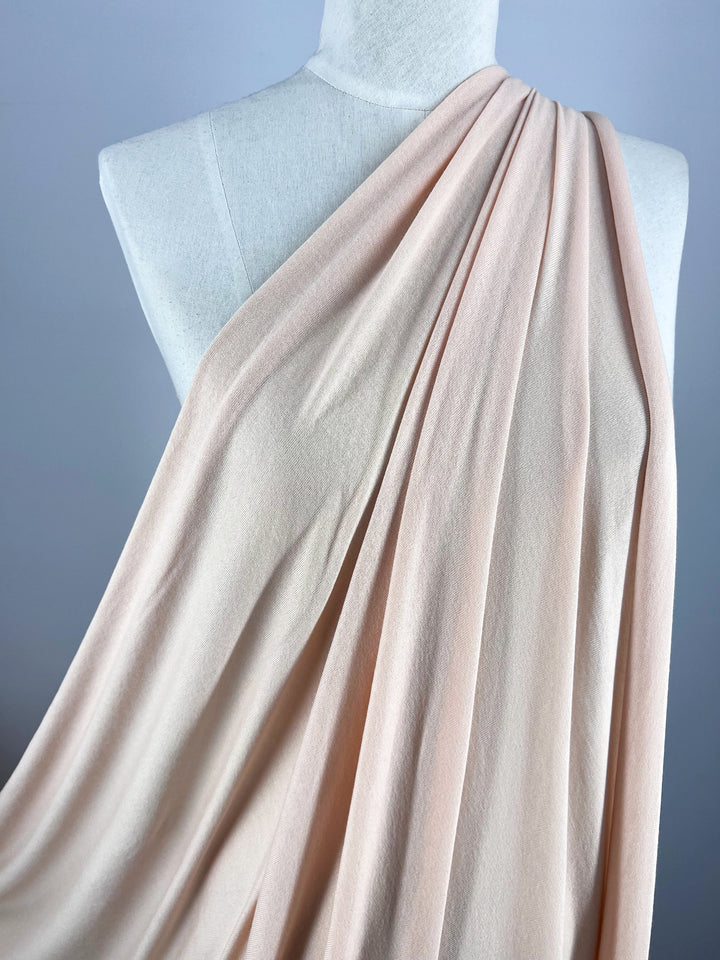 A mannequin features Super Cheap Fabrics' Summer Cotton Knit in Cream Tan (135cm), with lightweight, flowing cotton elegantly draping from one shoulder, creating gentle folds and a graceful appearance against the neutral backdrop.