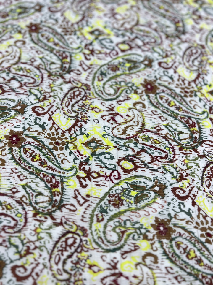View the intricate paisley pattern on the lightweight Voile - Paisley - 145cm cotton polyester fabric by Super Cheap Fabrics. Featuring green, brown, and yellow swirls on a white background, it's perfect for warm weather clothing.