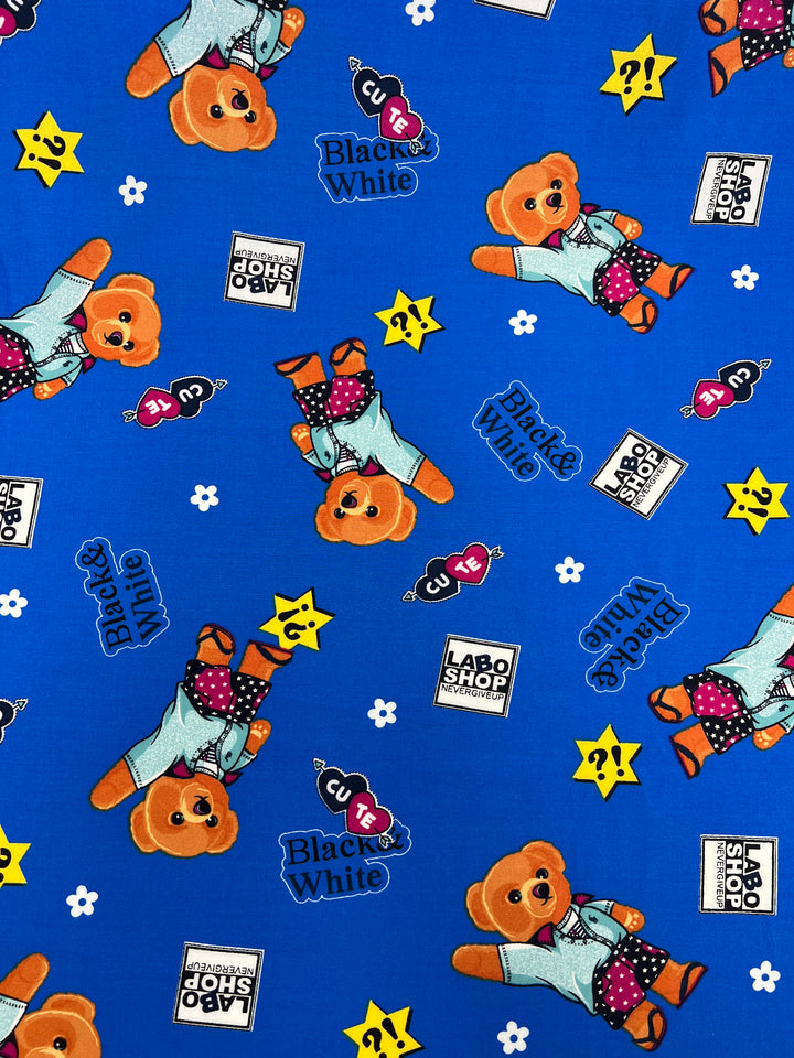 A blue fabric features a pattern with orange teddy bears in various poses wearing blue outfits. The design includes scattered text reading "Black & White" and "LABO SHOP" along with images of stars, keys, and question marks. Ideal for dresses, this Printed Rayon - Royal Bear - 145cm by Super Cheap Fabrics is both playful and stylish.