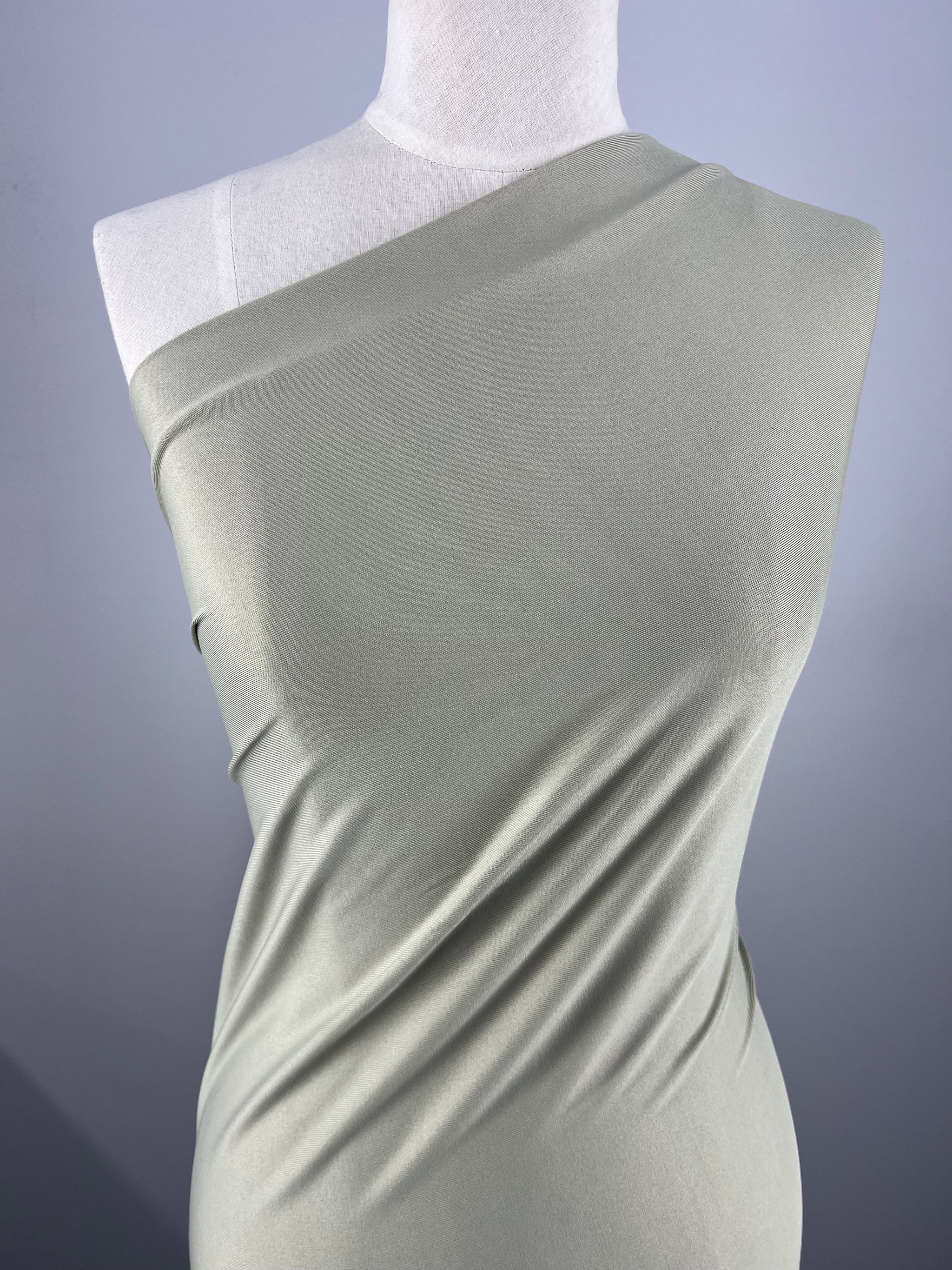 A minimalist dress form draped with a light olive green fabric, arranged asymmetrically on one shoulder, showcases the smooth, form-fitting qualities of 