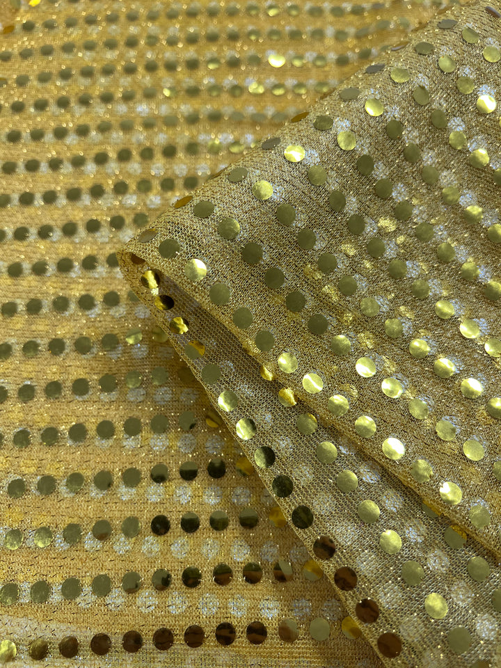 A close-up of Super Cheap Fabrics' Sequin - Gold - 112cm shows rows of round gold metallic sequins sewn onto sheer mesh, with part of the fabric folded over itself.