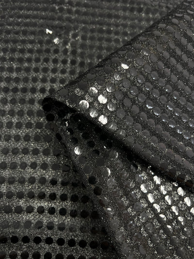 Close-up of Super Cheap Fabrics’ Sequin - Black - 112cm: shiny black polyester fabric with circular sequins in a grid pattern, shown folded over a flat background.