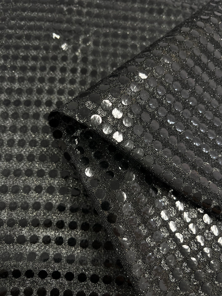 Close-up of Super Cheap Fabrics’ Sequin - Black - 112cm: shiny black polyester fabric with circular sequins in a grid pattern, shown folded over a flat background.