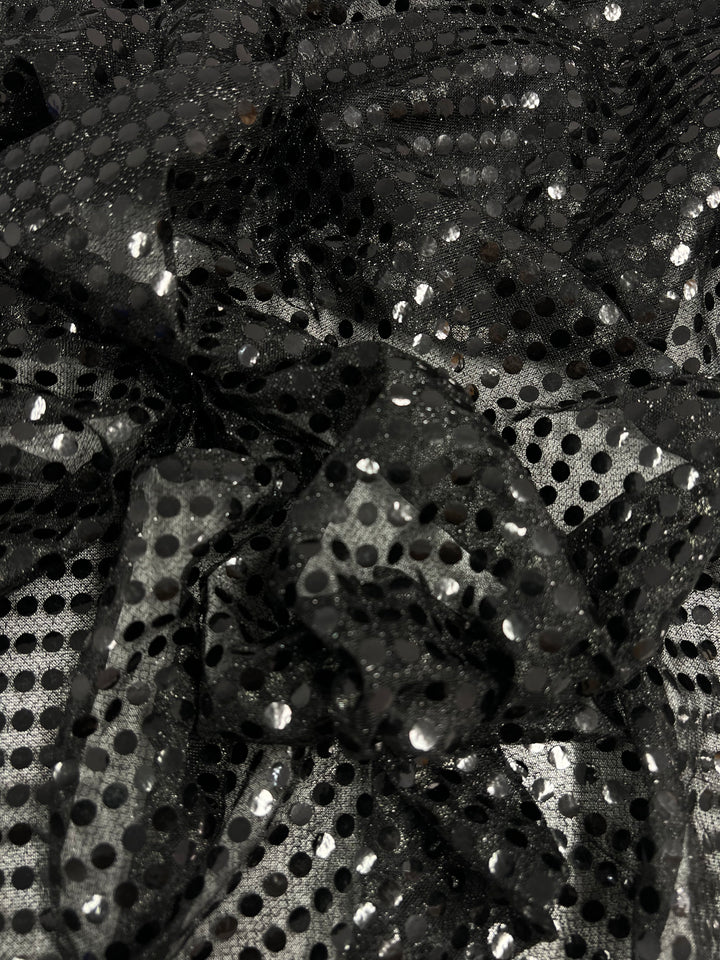 A close-up of Sequin - Black - 112cm from Super Cheap Fabrics shows crumpled lightweight polyester mesh, featuring shiny black sequins and polka dots that reflect light for a textured, glossy look.