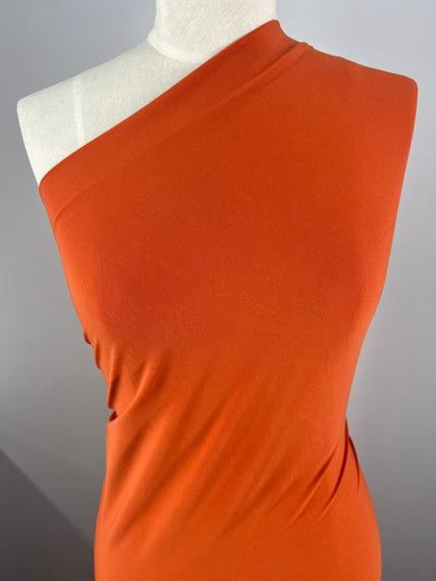 A mannequin is draped in a vibrant Super Cheap Fabrics ITY Knit - Burnt Orange - 150cm fabric. The polyester-spandex blend has a smooth texture and is wrapped tightly around the torso, highlighting the contours of the mannequin. The background is plain and gray, allowing the fabric's color to stand out.