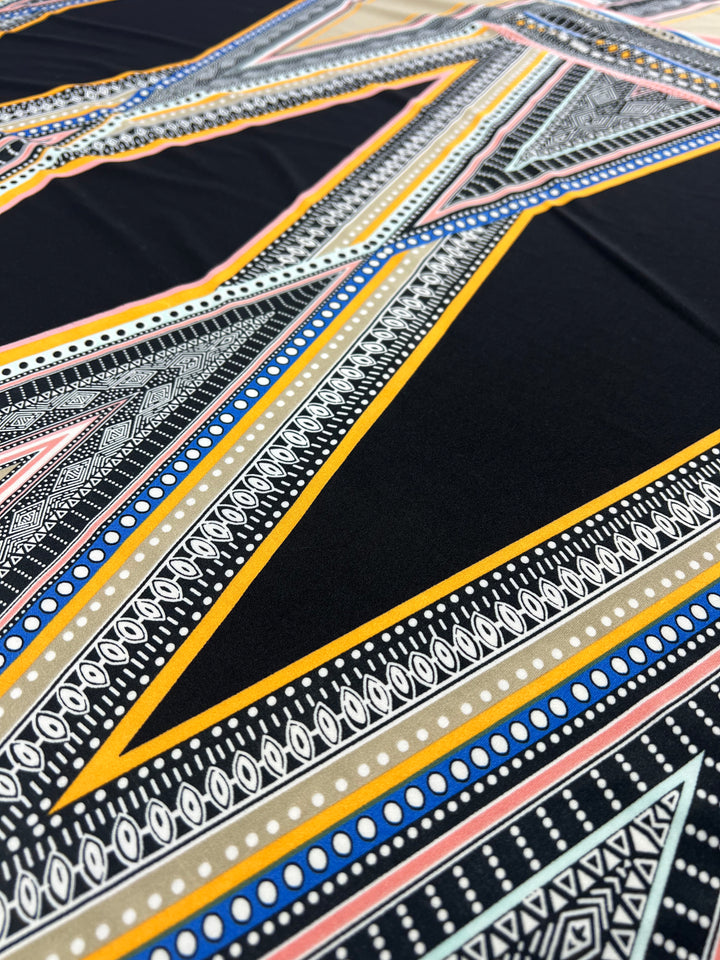 The REMNANT - Lycra - Aztec Wrap from Super Cheap Fabrics showcases a close-up of its 160cm panel, featuring intricate geometric patterns. The design incorporates zigzag lines, dots, and triangles in shades of blue, yellow, black, and white for a bold and vibrant visual impact.