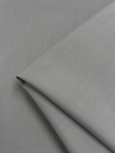 A close-up of Poplin - Mirage Grey - 112cm by Super Cheap Fabrics shows neatly folded, light gray poly cotton fabric with a smooth, slightly textured surface, perfect for childrenswear, on a matching gray background.