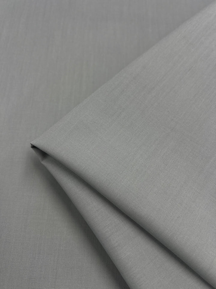 A close-up of Poplin - Mirage Grey - 112cm by Super Cheap Fabrics shows neatly folded, light gray poly cotton fabric with a smooth, slightly textured surface, perfect for childrenswear, on a matching gray background.