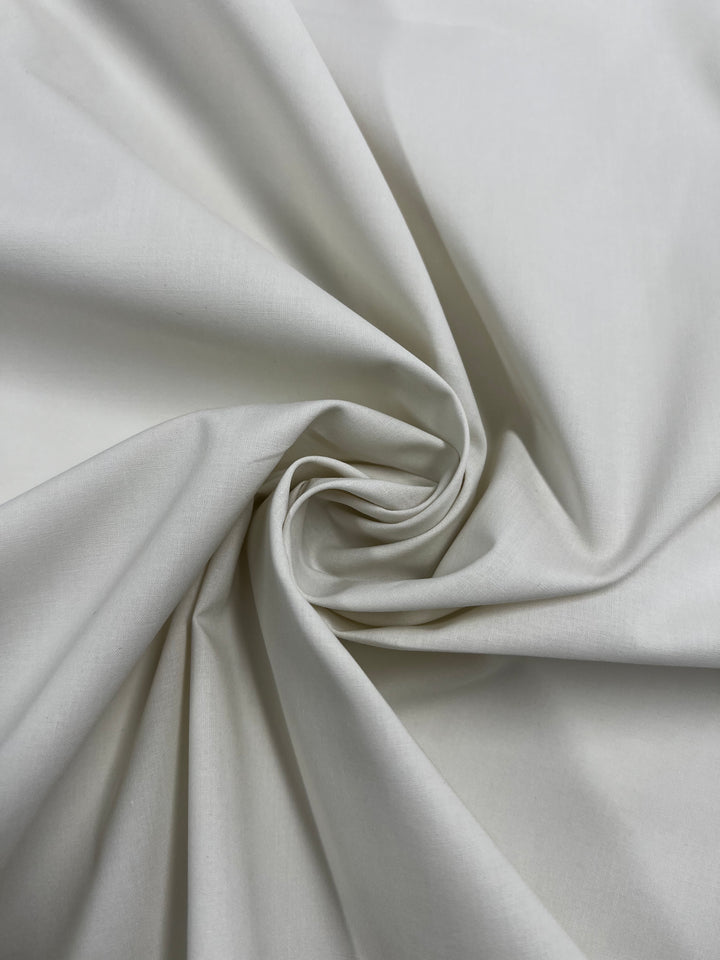 A close-up of Super Cheap Fabrics' Plain Cotton - Birch - 150cm shows soft, light beige 100% cotton arranged in gentle folds that spiral inward, highlighting its smooth texture and subtle swirling pattern.