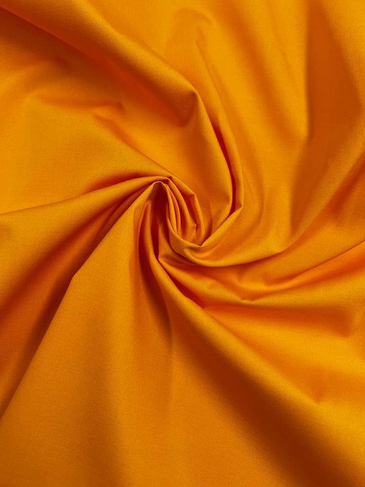 Super Cheap Fabrics' Plain Cotton - Zinnia - 147cm is a lightweight, 100% cotton fabric in a vibrant orange shade with a subtle sheen, arranged in soft spiral folds that showcase its smooth texture and vivid color.