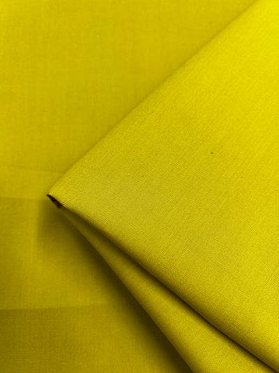 A close-up of Super Cheap Fabrics' Plain Cotton - Antique Moss (150cm), a lightweight, smooth 100% cotton in a rich antique moss yellow, partially folded on a flat surface and draping beautifully.